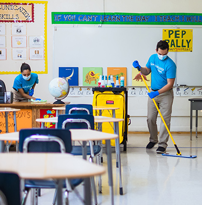 Two cleaning people are cleaning the inside of a classroom by wiping down desks and using a microfiber mop to wipe dust and debris off floors